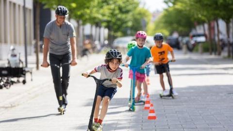 Eine Gruppe Kinder sowie ein Erwachsener fahren auf Tretrollern um Verkehrshüchen