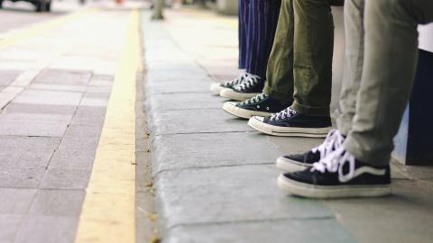 Auf einem Gehweg sind mehrere Schuhe und Beine von Menschen zu sehen, diese sitzen offensichtlich auf einer Bank. Links ist die Straße mit einer gelben Markierung zu sehen.