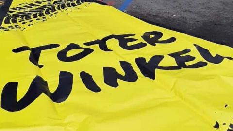 Eine gelbe Plane liegt auf dem Boden. Die Aufschrift "Toter Winkel" ist zu lesen, im oberen Bildrand sind Reifenabdrücke zu sehen
