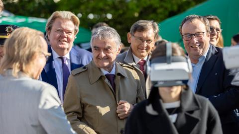 Die beiden NRW-Minister Herbert Reul und Oliver Krischer stehen an einem Stand und sprechen mit einem Mann. Unten im Bild ein Kind mit einer großen Computer-Brille auf