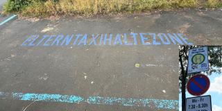 Auf einer Straße ist in blauer Farbe der Schriftzug "Elterntaxihaltezone" angebracht. Rechts im Bild ist das dazugehörige Straßenschild eingeblendet